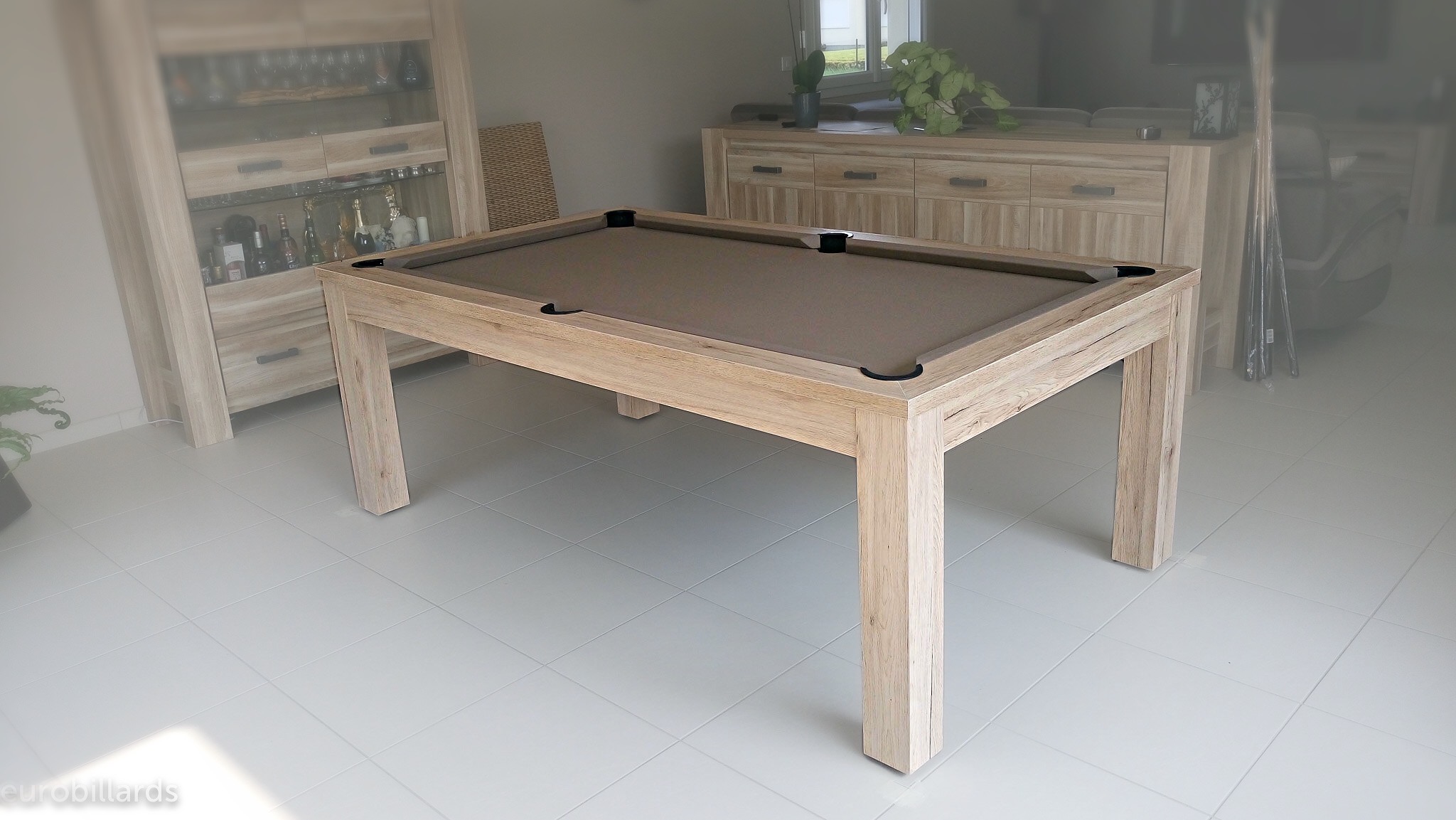 billard trendy en chêne éclairci veiné avec son tissu taupe trone sur le carrelage blanc du salon entouré du mobilier en chêne clair dans le salon derrière la salle à manger